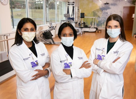 Photo of three physiatrists standing with their arms crossed in a clinical setting, wearing white coats and masks